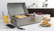 Compact stainless steel electric grill cooking breakfast foods including pancakes, eggs, bacon, and sausage on a marble countertop, surrounded by orange juice, coffee mugs, fresh berries, and a modern kitchen backdrop.