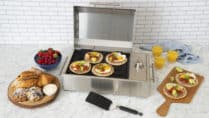 Compact stainless steel electric grill cooking breakfast tacos topped with eggs and avocado on a marble countertop, surrounded by pastries, fresh berries, and orange juice in a bright modern kitchen setting.