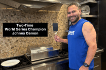 Former professional baseball player Johnny Damon standing in a luxury kitchen beside a compact stainless steel electric grill, holding a spatula with promotional text highlighting his two-time World Series Championship title.