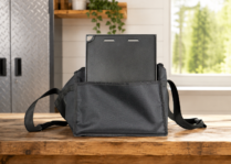 Alt text: Black electric grill carry bag with a flat grill component inside, placed on a wooden workbench in a clean workshop-style setting with metal cabinets, a plant shelf, and a bright window in the background.