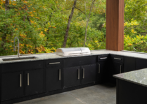 Built-in stainless steel electric grill in a modern outdoor kitchen with black cabinetry, stone countertops, a sink, and a lush wooded backdrop.