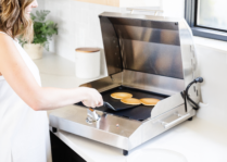 Smiling woman cooking pancakes on a stainless steel Kenyon electric griddle in a bright modern kitchen.