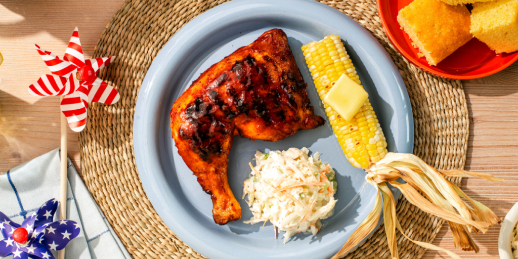 Overhead view of a Kenyon electric grill cooking two barbecue chicken leg quarters with grill marks, alongside a plated chicken dinner with corn on the cob, coleslaw, cornbread, and patriotic table décor.