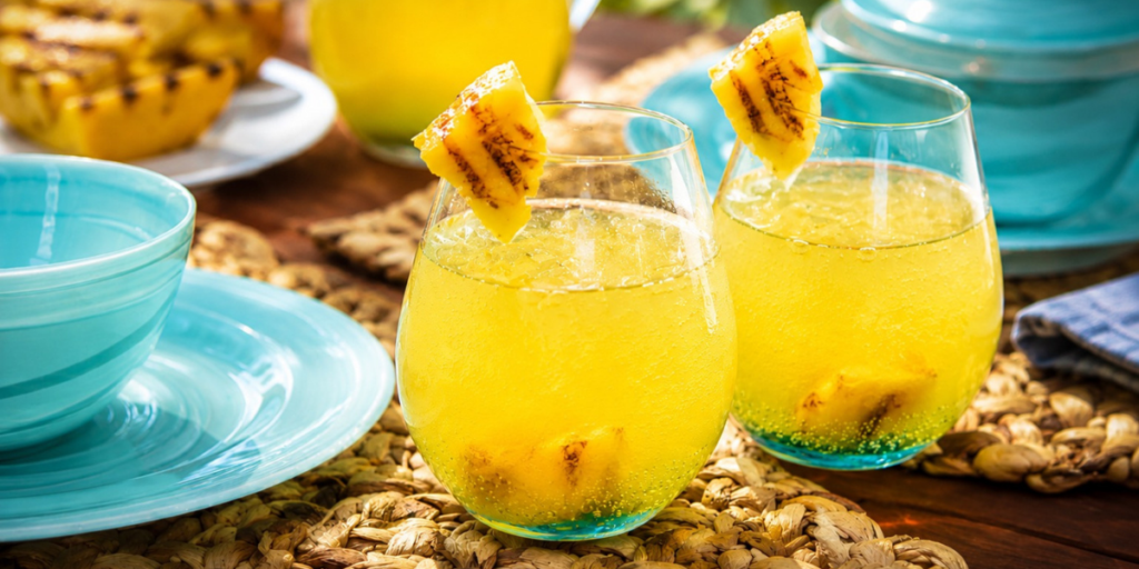 Two glasses of grilled pineapple lemonade served over ice with pineapple chunks and grilled pineapple garnishes, set on a table with tropical dinnerware and grilled pineapple in the background.