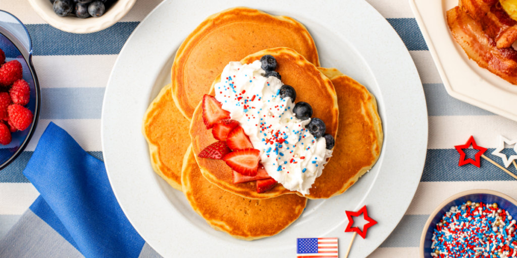 Overhead view of a stack of pancakes topped with whipped cream, strawberries, blueberries, and red, white, and blue sprinkles, served with bacon, scrambled eggs, fresh berries, and maple syrup on a festive breakfast table