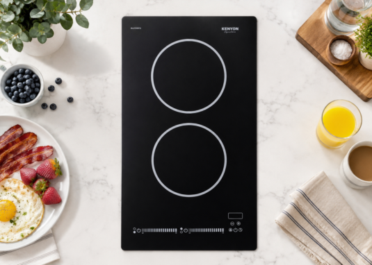 Overhead view of a Kenyon Signature two-burner induction cooktop installed in a white marble countertop, surrounded by breakfast items, greenery, coffee, orange juice, and kitchen linens.