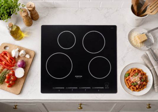 Overhead view of a Kenyon Signature black glass cooktop installed in a white marble countertop with fresh ingredients and kitchen styling arranged around it.