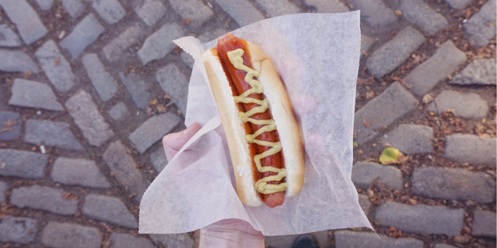 Hand holding a classic New York–style hot dog wrapped in deli paper, topped with spicy brown mustard and served in a soft bun. The hot dog is photographed from above against a cobblestone street background, evoking a traditional New York City street-food scene.