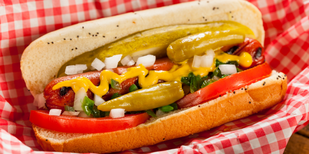 Chicago-style hot dog with a grilled all-beef frankfurter topped with yellow mustard, chopped white onions, bright green relish, tomato wedges, a dill pickle spear, and sport peppers, served in a poppy seed bun on red-and-white checkered paper.