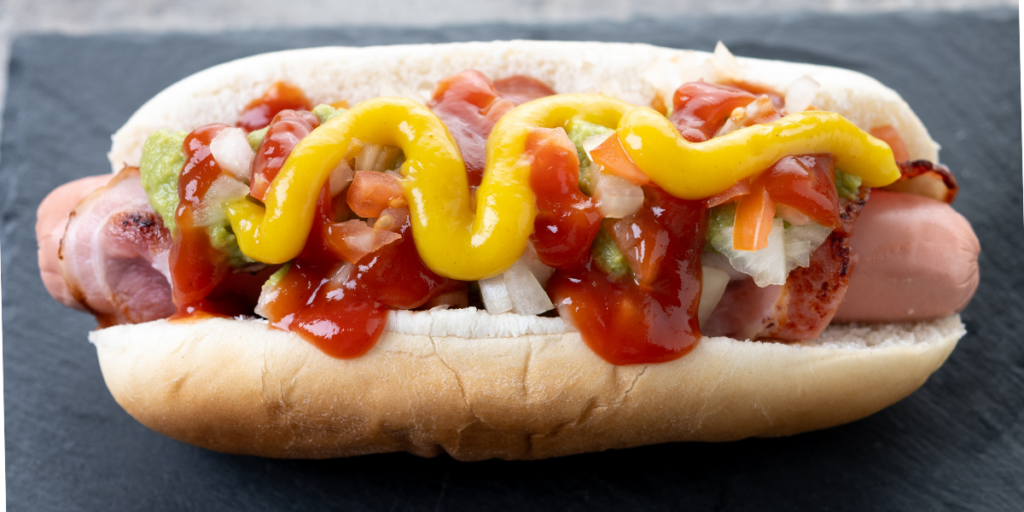 Sonoran-style hot dog wrapped in bacon and topped with diced onions, fresh tomatoes, green salsa, ketchup, and yellow mustard, served in a soft bun on a dark serving surface.