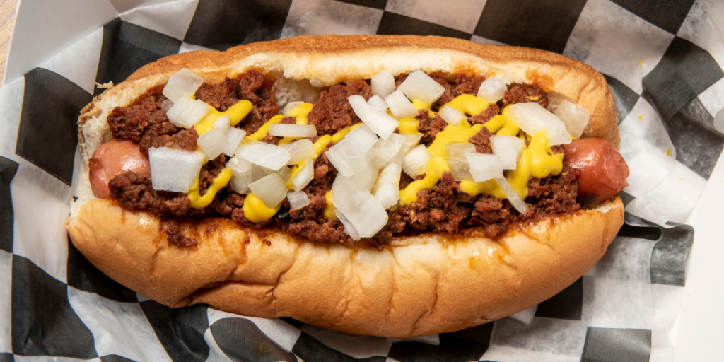 Detroit-style Coney dog topped with savory meat chili, yellow mustard, and diced white onions, served in a steamed bun on black-and-white checkered deli paper.