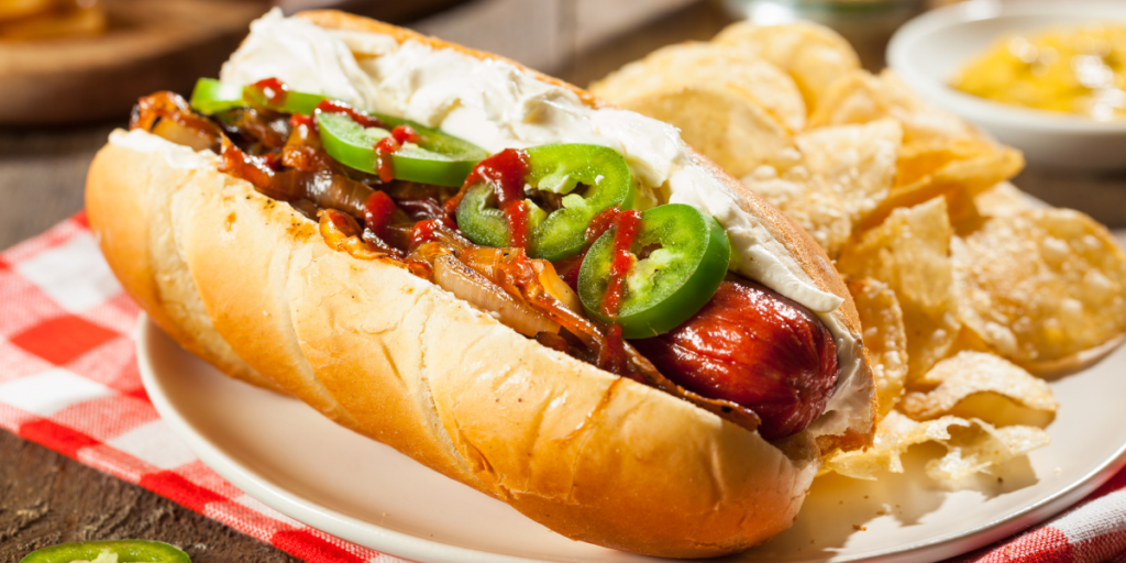 Seattle-style hot dog topped with cream cheese, grilled onions, sliced jalapeños, and a drizzle of sauce, served in a toasted bun alongside potato chips.