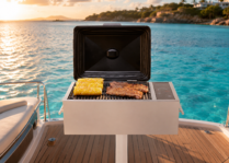 Stainless-steel electric grill mounted on the aft deck of a luxury yacht, grilling corn on the cob and sliced steak with the lid open. The grill is positioned against a backdrop of expansive Caribbean-blue ocean under golden-hour sunlight. Gentle waves reflect warm sunlight, while a distant rocky coastline with upscale waterfront homes is softly blurred in the background. Minimal boat structure is visible, emphasizing the open water and luxury boating lifestyle. The scene has an editorial, high-end outdoor living aesthetic with the grill as the primary focal point.