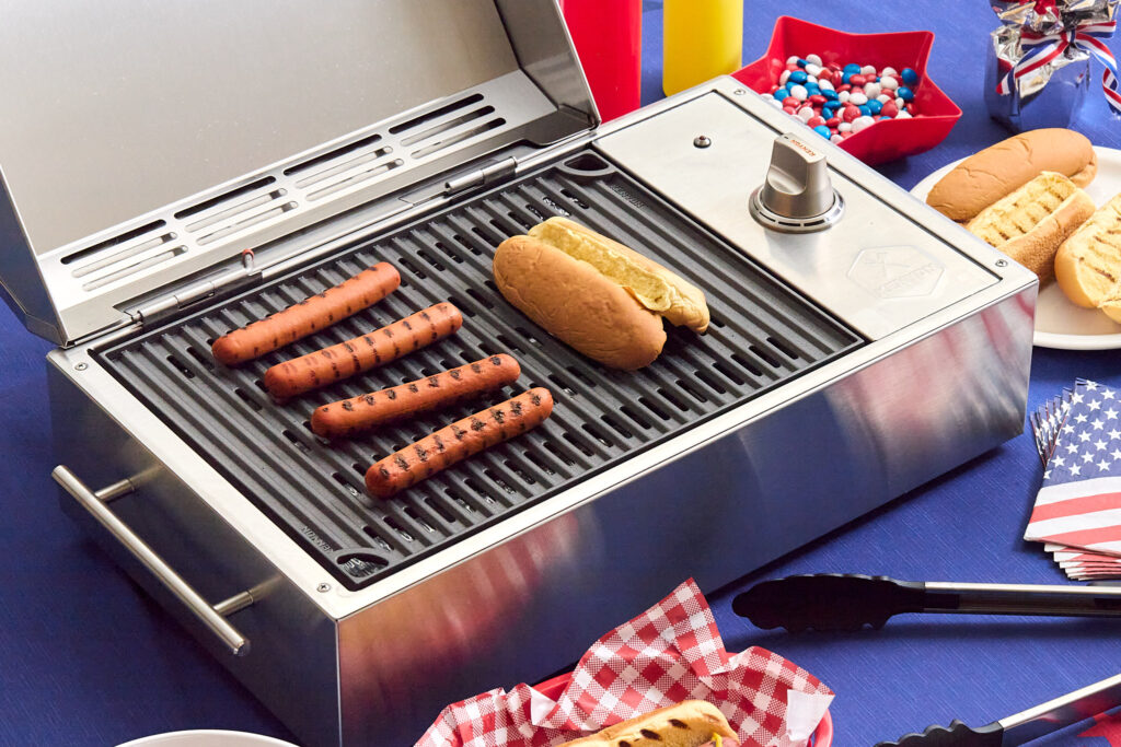 Kenyon City Grill cooking hot dogs and toasting buns on a stainless steel electric grill, surrounded by patriotic red, white, and blue decorations for a summer cookout.