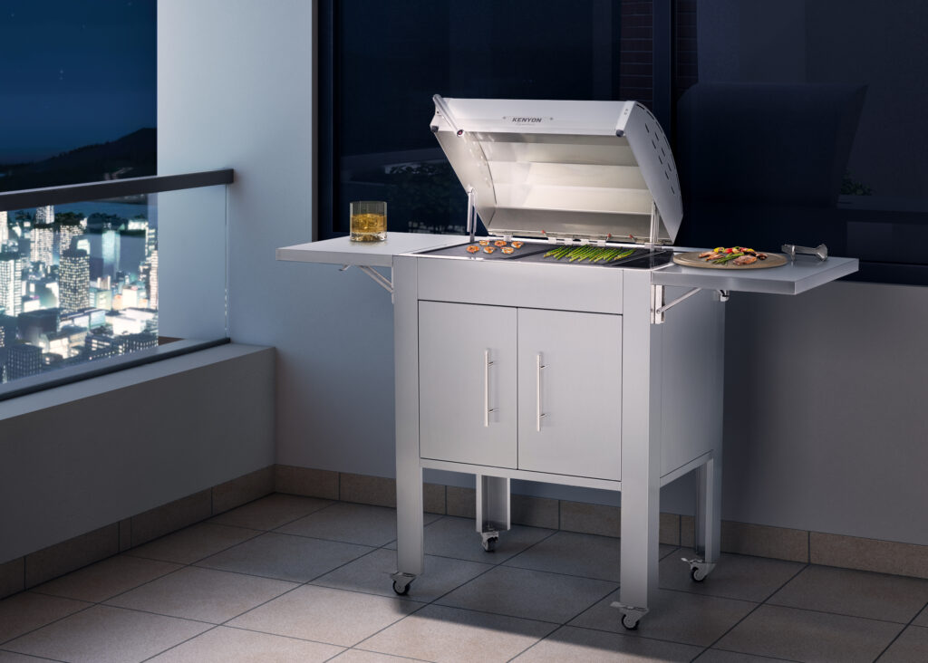 Kenyon Signature electric grill with a white lid and stainless steel cart on a modern balcony at night. The lid is open, revealing illuminated grill grates cooking asparagus and shrimp, with city lights visible in the background.
