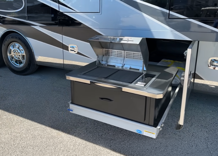 Kenyon Texan electric grill built into a slide-out outdoor kitchen on a motorhome, with the stainless steel lid open and grilling surface visible.