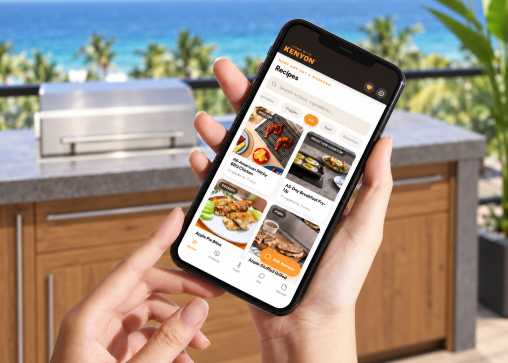 Hands holding a phone displaying the Kenyon app’s recipe collection, with an outdoor grill and ocean view in the background.