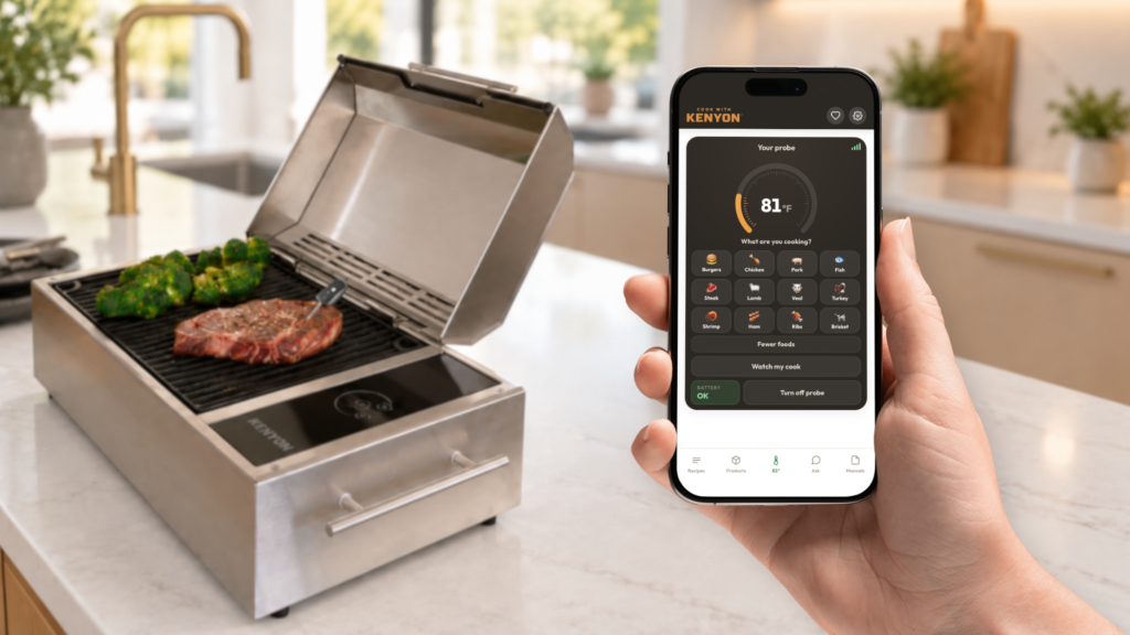 Hand holding a phone displaying a probe temperature of 81°F in the Kenyon app beside a countertop grill cooking steak and broccoli.